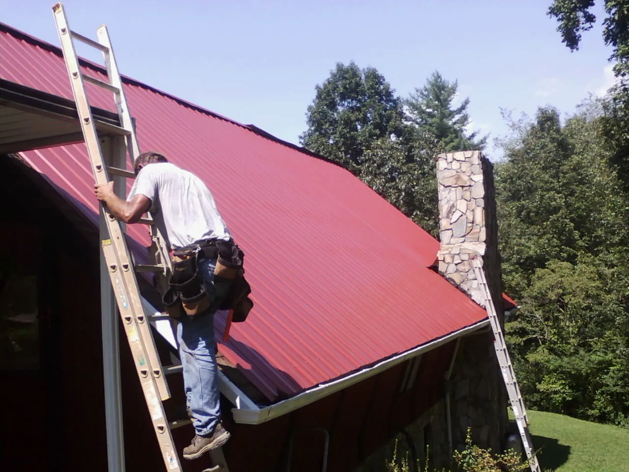 Roofer on a ladder beside a red metal roof and stone chimney — Soffit & Fascia Repair by Roofing Brothers in Bowie